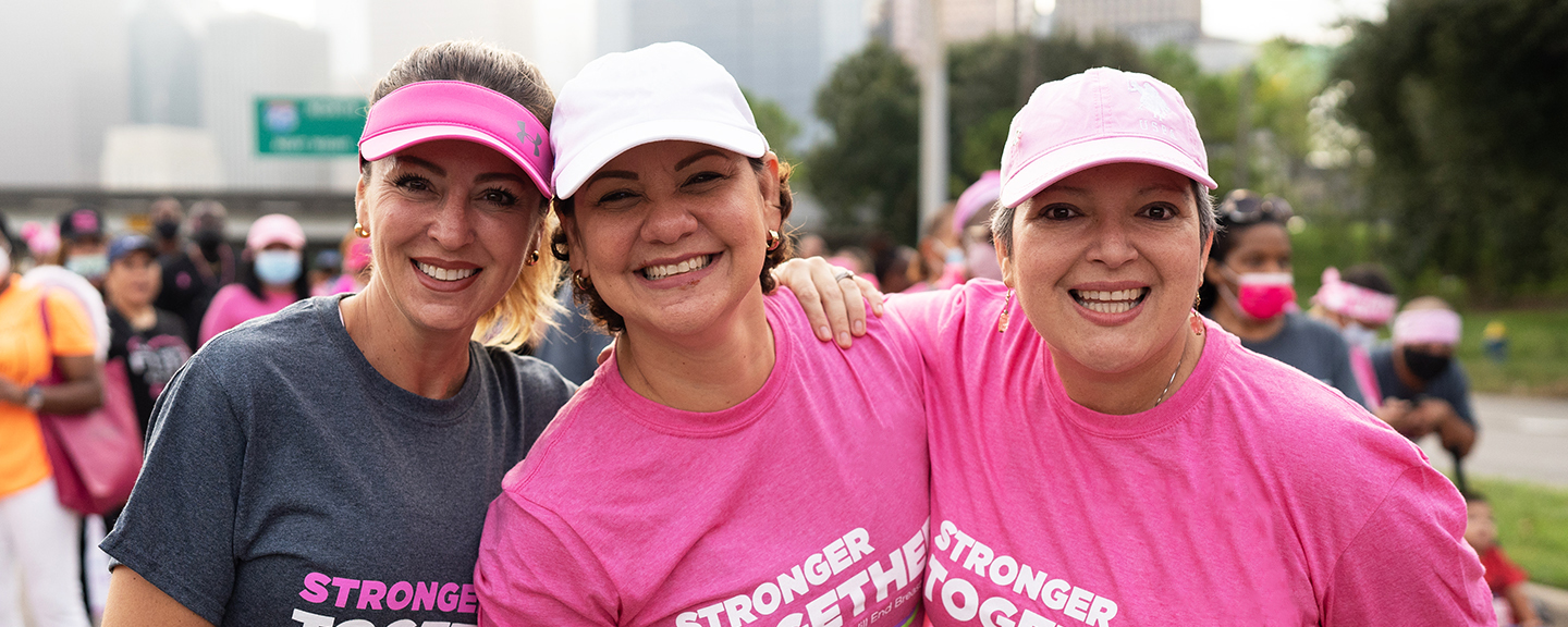 Three women in pink shirts smiling in front of a crowd before a Susan G. Komen walk. 