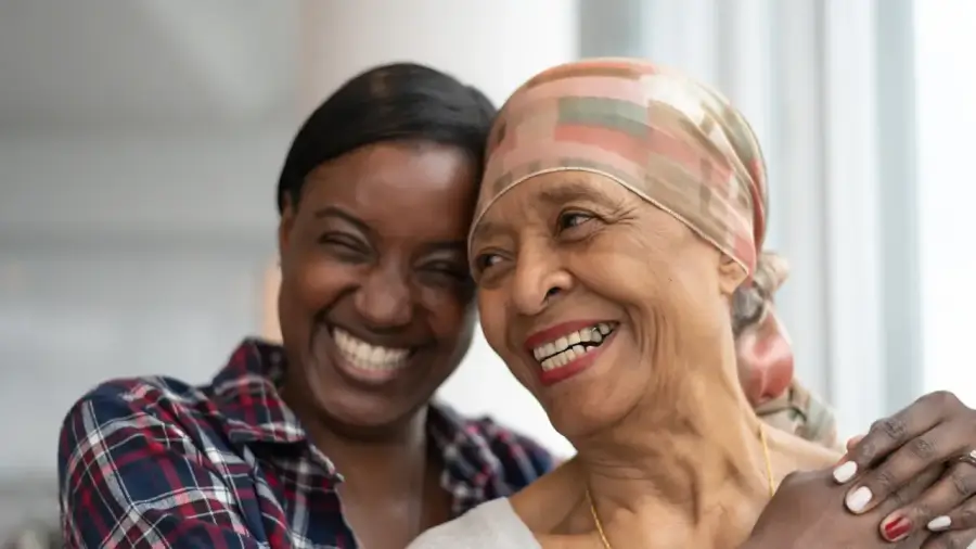 A senior African American female cancer patient smiles while a female family member hugs her.