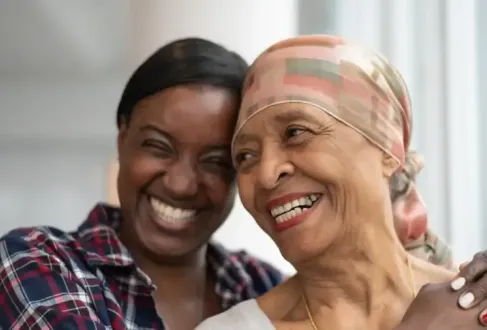 A senior African American female cancer patient smiles while a female family member hugs her.